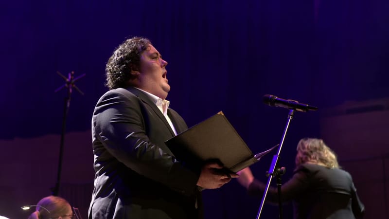 A soloist in formal attire holds a black folder while singing at the microphone during a performance.