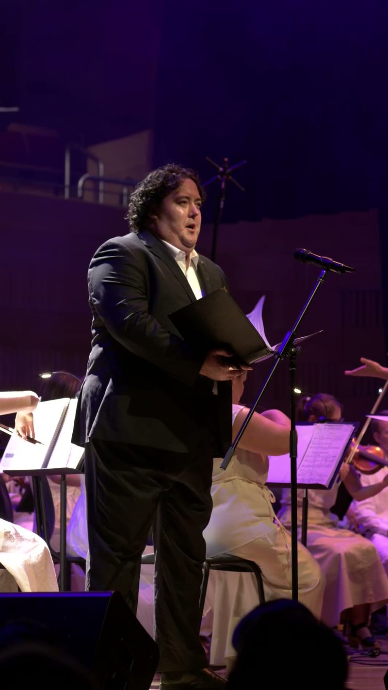 A soloist in formal attire holds a folder while performing at the microphone with orchestra members visible in the background.