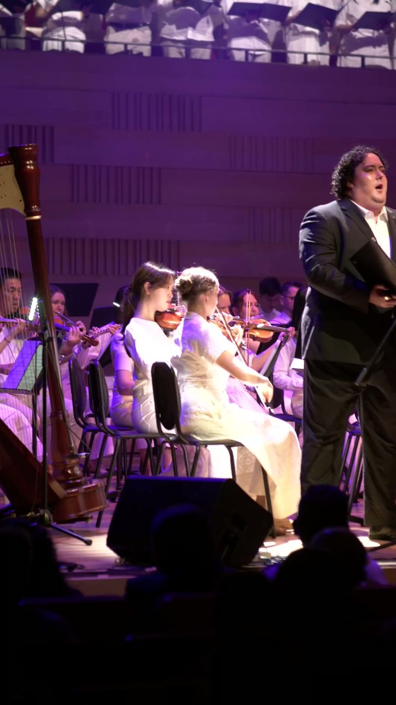 A male tenor soloist performs with young string players and orchestra members in formal concert attire under purple stage lighting.