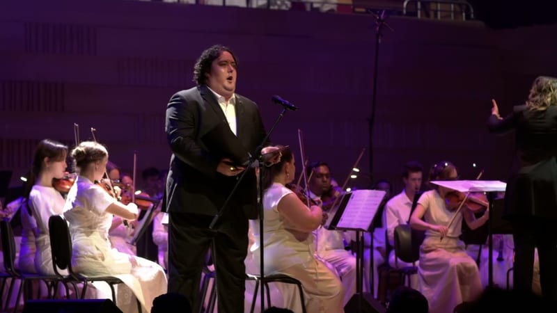 A male soloist in formal attire performs with an orchestra and choir members dressed in white, illuminated by purple stage lighting.