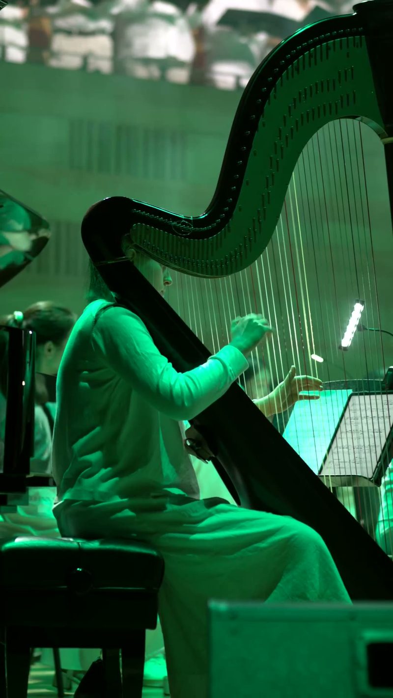 A harpist performs at a concert, illuminated by green stage lighting as they play their instrument.