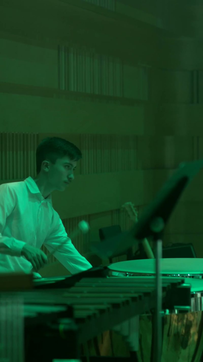 A young musician in a light-colored shirt plays drums during an orchestral performance in green-tinted lighting.