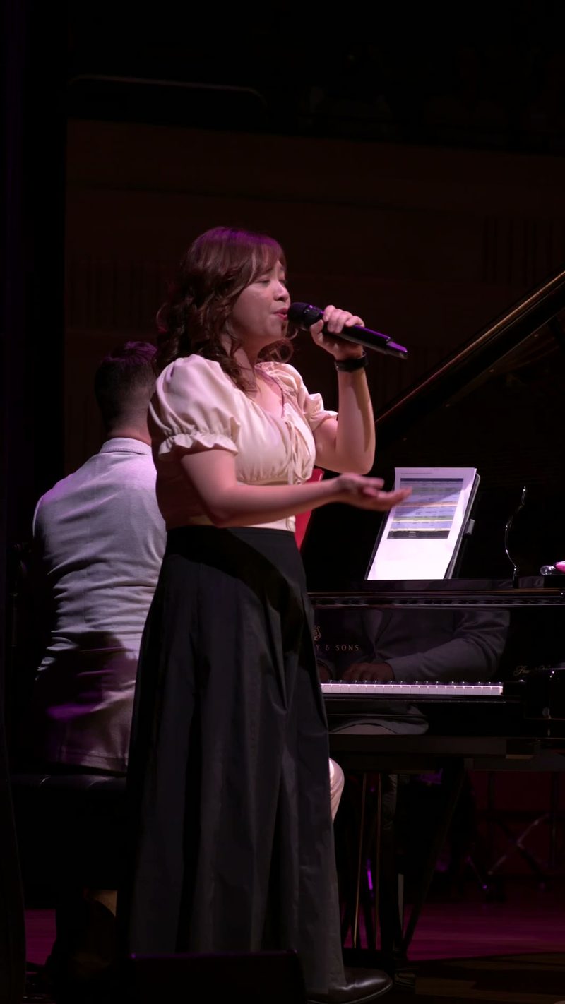 A female soloist performs at the microphone with pianist accompaniment on stage under purple lighting.