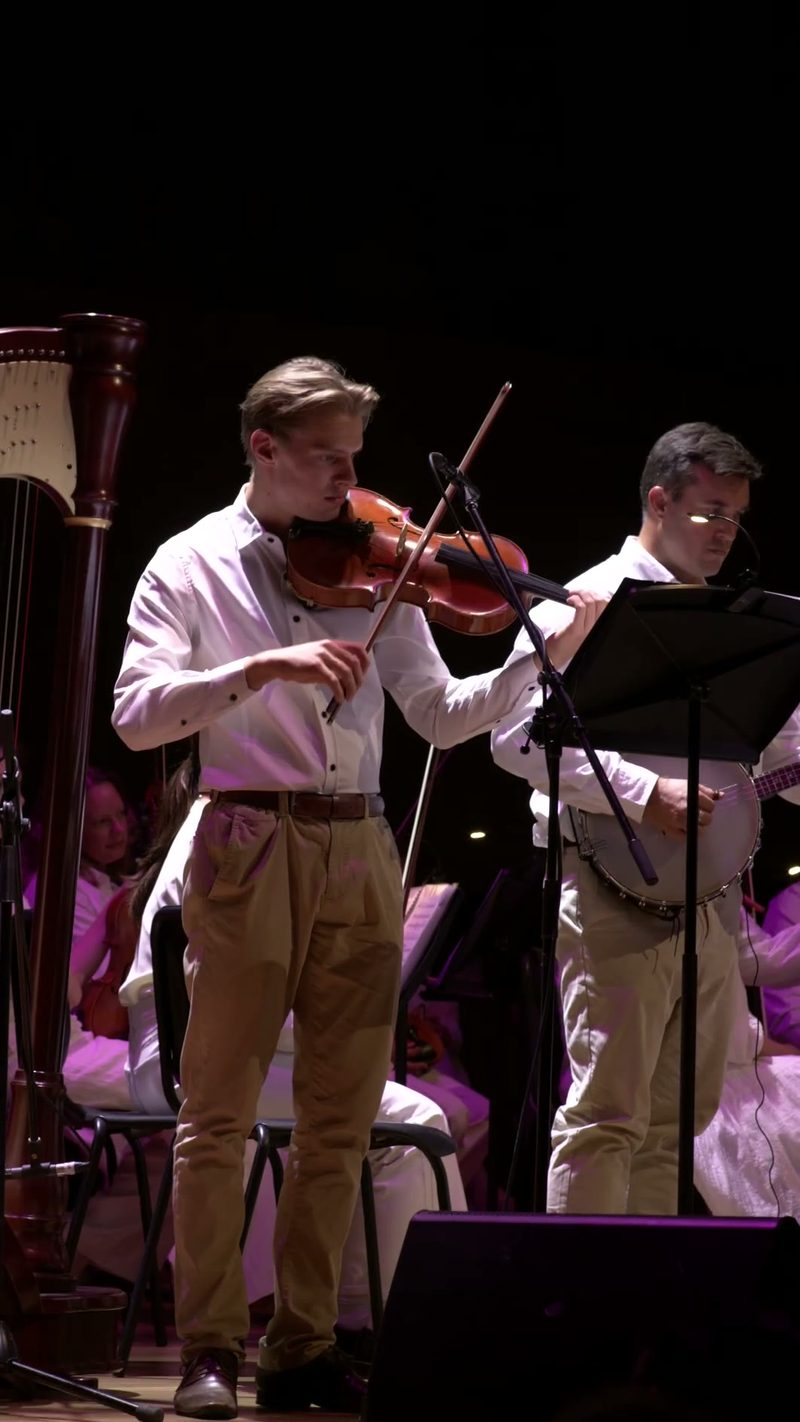 Young violinist performs with pianist during an orchestral concert under purple stage lighting.