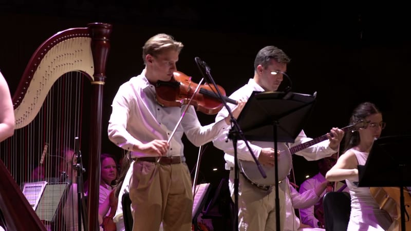 A violinist performs alongside a pianist and guitarist under purple stage lighting during an orchestral performance.