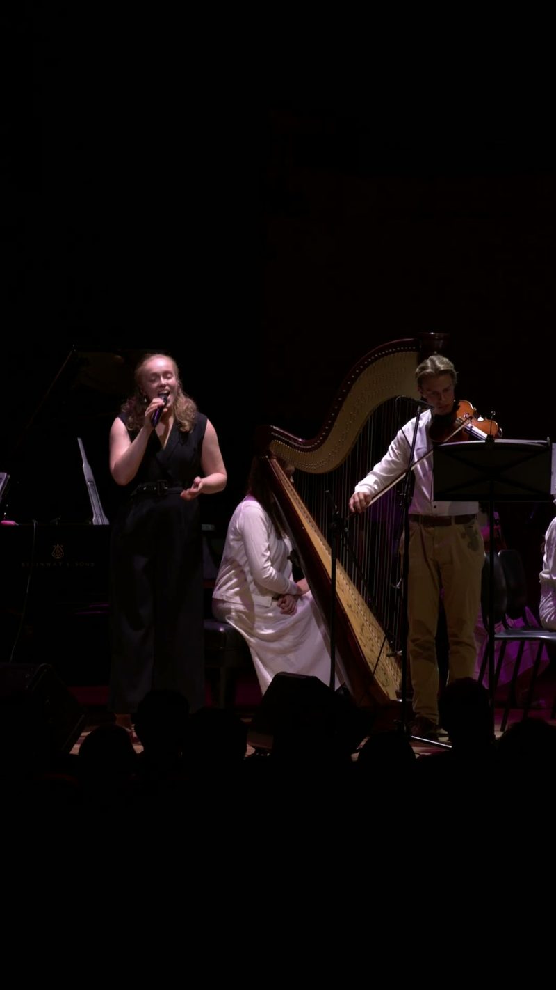 A female vocalist performs with a harpist and ensemble on stage during an orchestral concert.