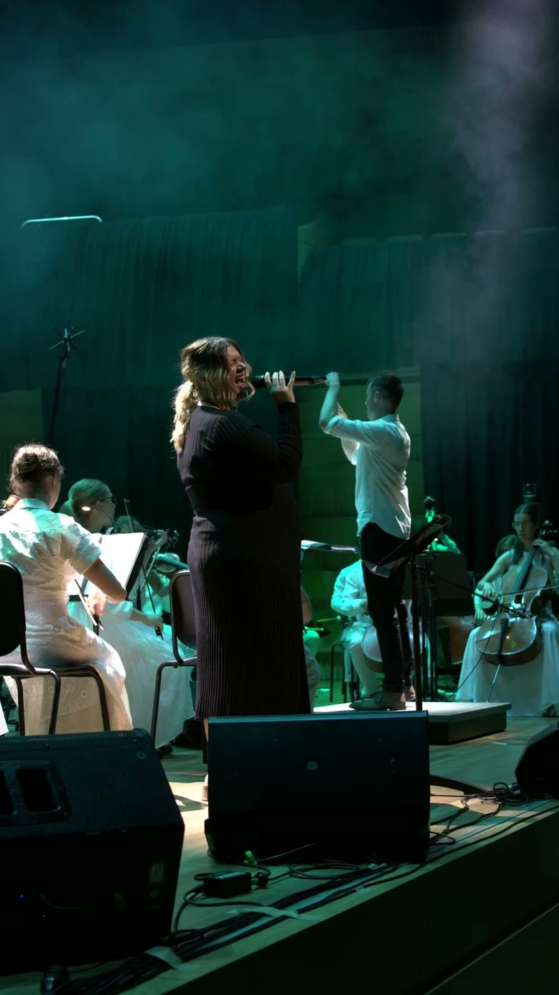 A female soloist performs with a flute while accompanied by an orchestra and choir on stage.