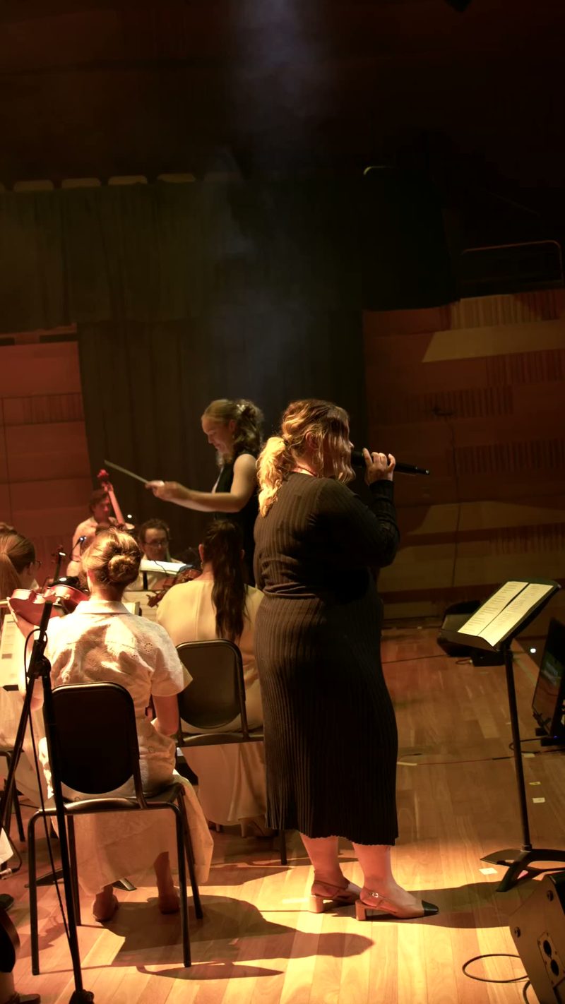 A conductor leads young singers during a choral performance on stage with warm theatrical lighting.