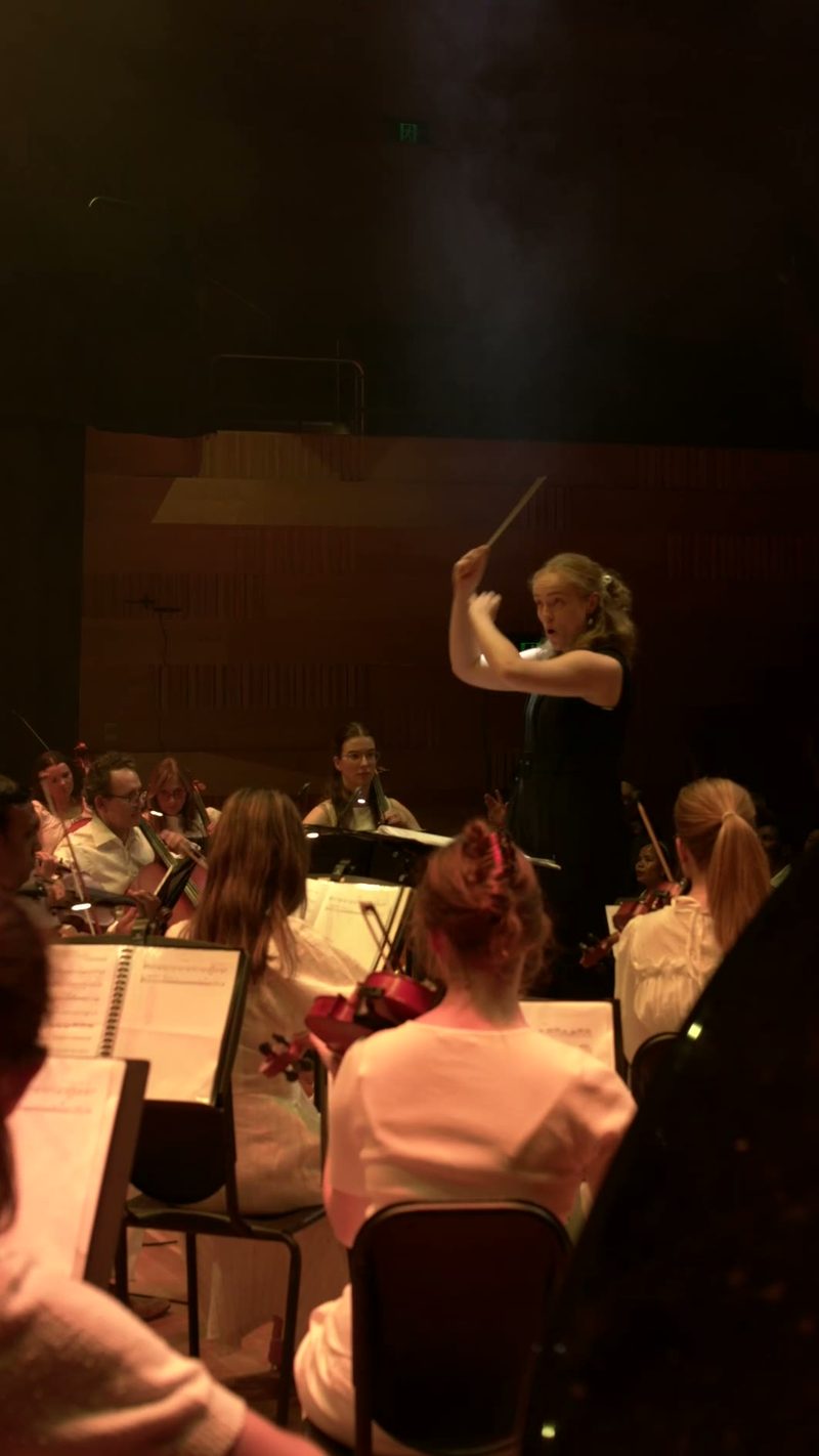 A female conductor leads an orchestra during a rehearsal or performance, with string and wind players visible in the ensemble.