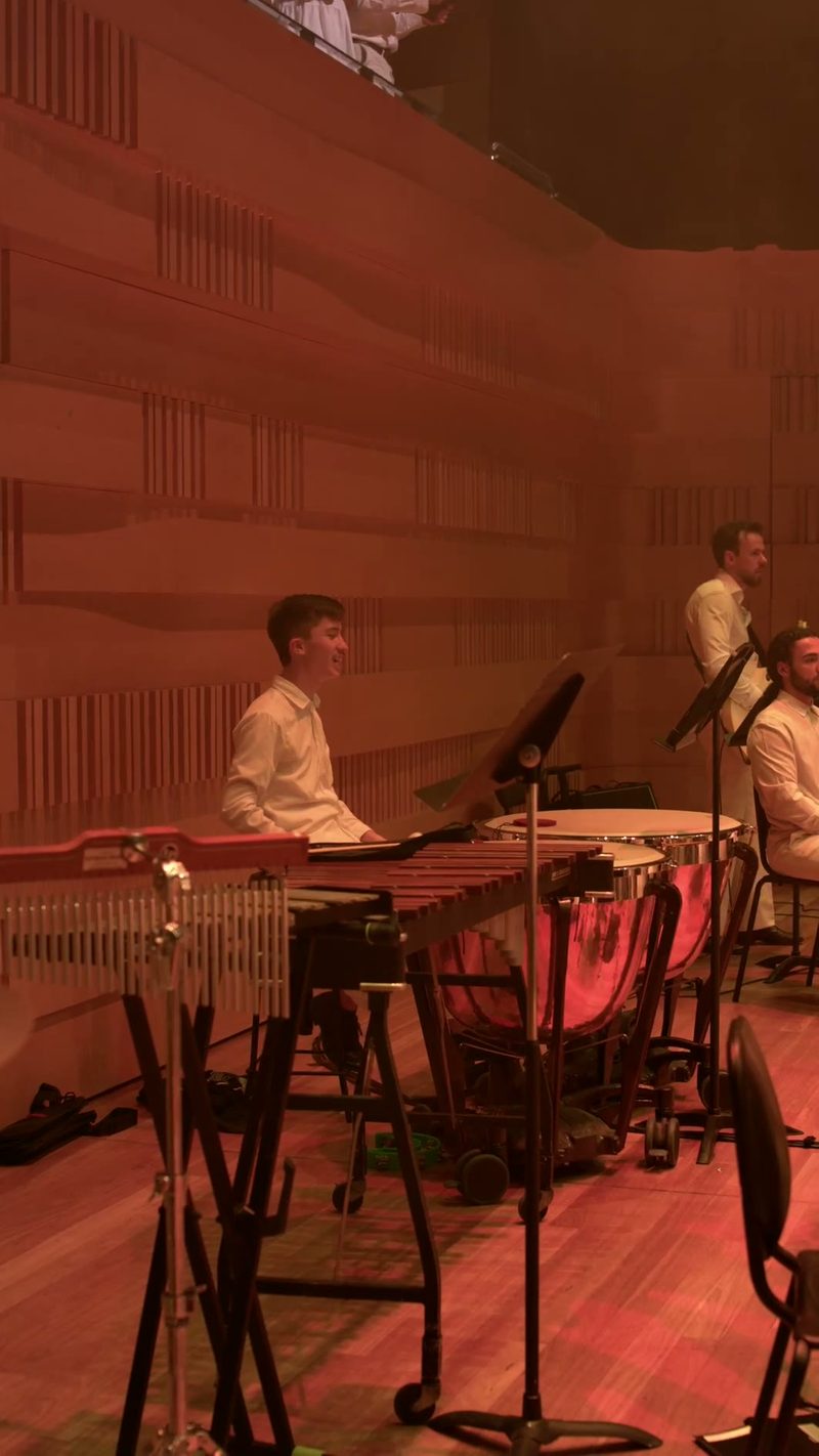Three musicians perform on percussion instruments including vibraphone and drums on stage with red ambient lighting.
