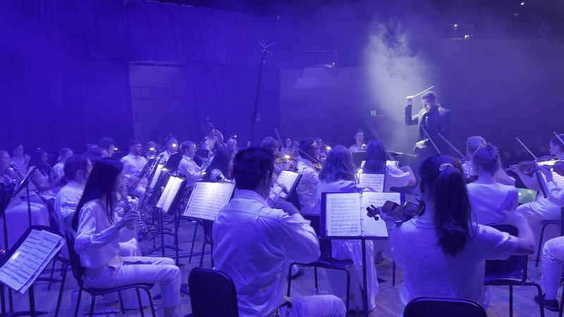 A conductor leads a large orchestra ensemble in a dramatic performance under vibrant purple stage lighting.