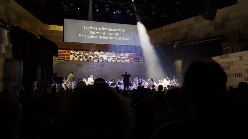 A full orchestra and choir perform together on stage with a projection displaying lyrics about resurrection and faith in Jesus.