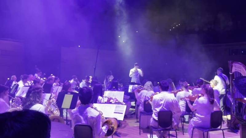 A conductor leads a full orchestra and choir ensemble under dramatic purple stage lighting during a symphonic performance.