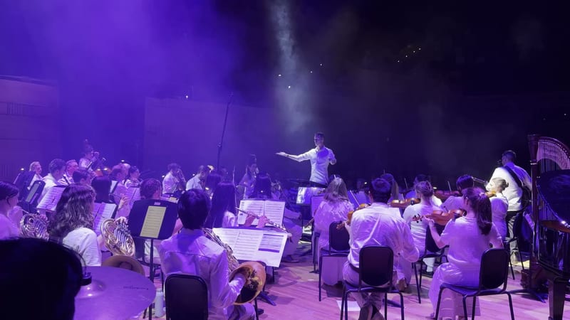 A conductor leads a large orchestra during a dramatic purple-lit performance on stage.