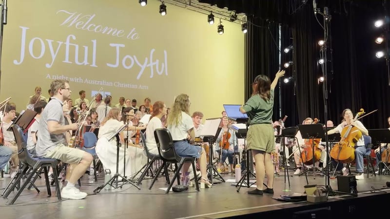 A conductor leads the Australian Christian Arts orchestra during a performance at the Joyful Joyful event, with string and wind players visible on stage.