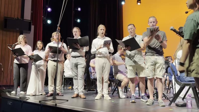 A choir performs together on stage, holding sheet music and singing in unison under stage lighting.