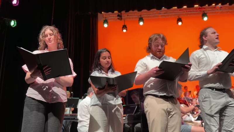 Four singers perform together on stage while reading from sheet music against an orange backdrop.