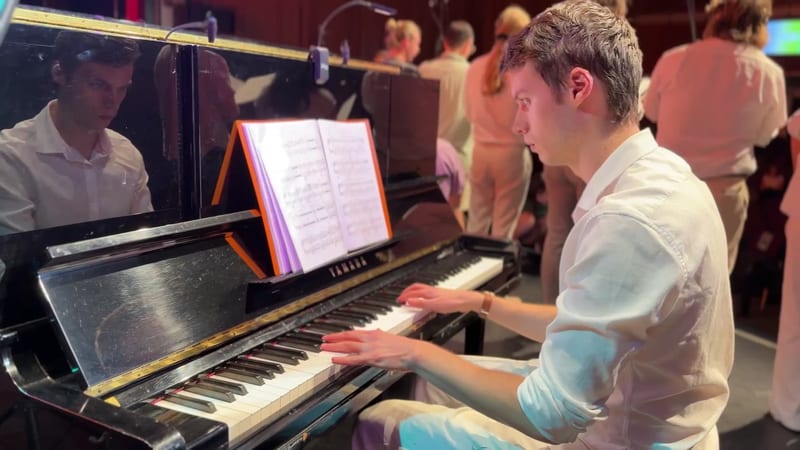 A young pianist performs at the grand piano while reading sheet music during an orchestral concert.