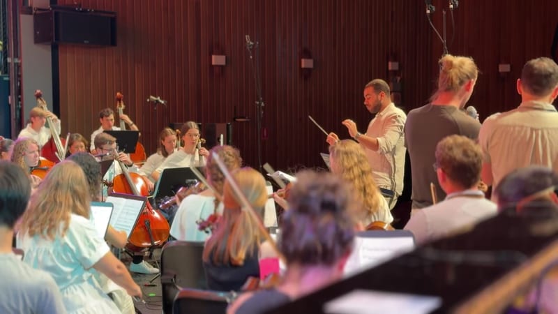A conductor leads a string orchestra section during a rehearsal in a professional concert hall.