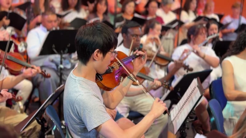 A young violinist performs with sheet music during an orchestral rehearsal or concert with an engaged audience in the background.