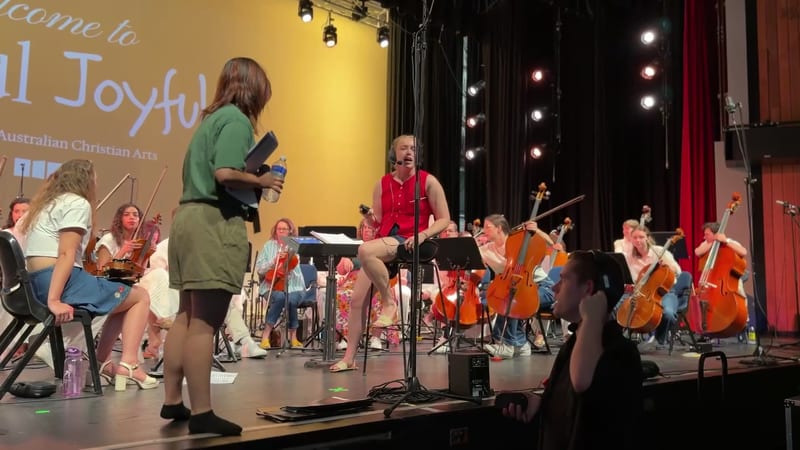 Young musicians from the orchestra perform during a rehearsal or concert at Australian Christian Arts, with string players seated and conductor engaging with performers.