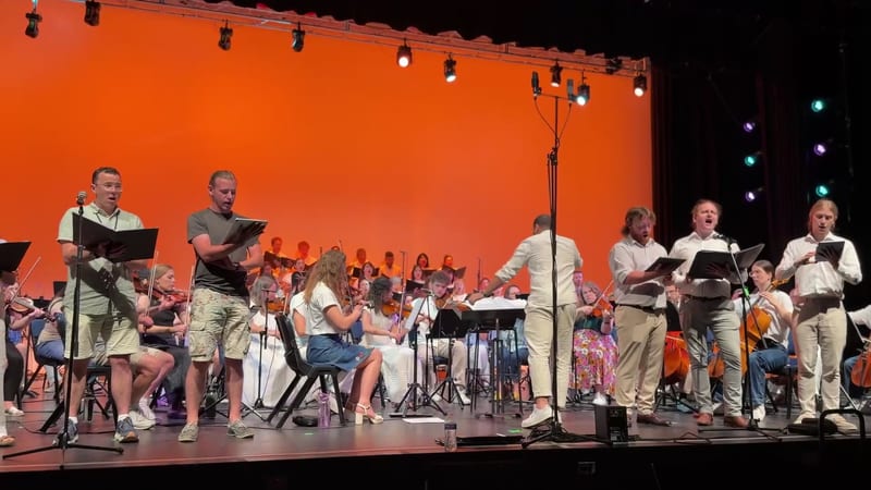 A mixed ensemble of singers and instrumentalists perform together on stage with an orchestra in the background under dramatic orange lighting.