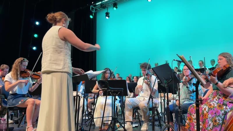 A conductor leads a large ensemble of string players and singers during a performance on stage with turquoise lighting.