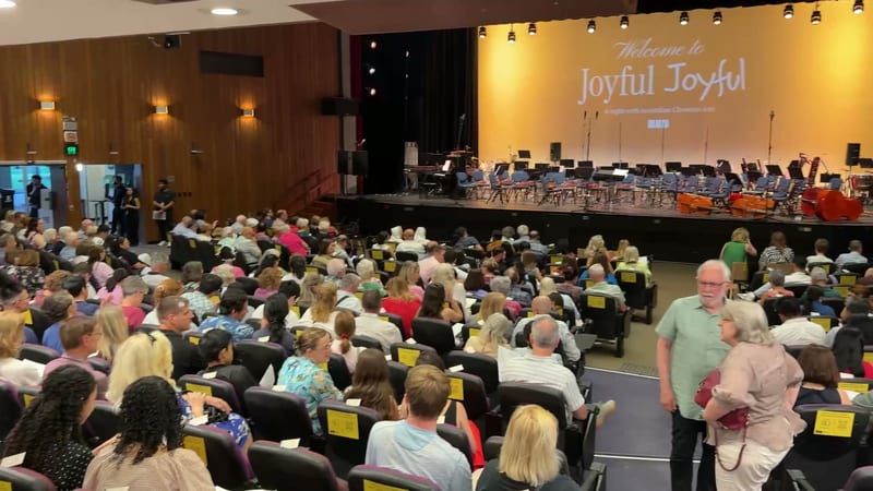 A large audience fills the auditorium for a Joyful Joyful concert event, with the orchestra and choir visible on stage.