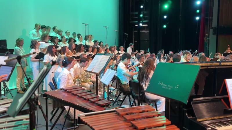 A youth orchestra and choir perform together on stage with percussion instruments in the foreground and singers positioned at elevated risers in the background.