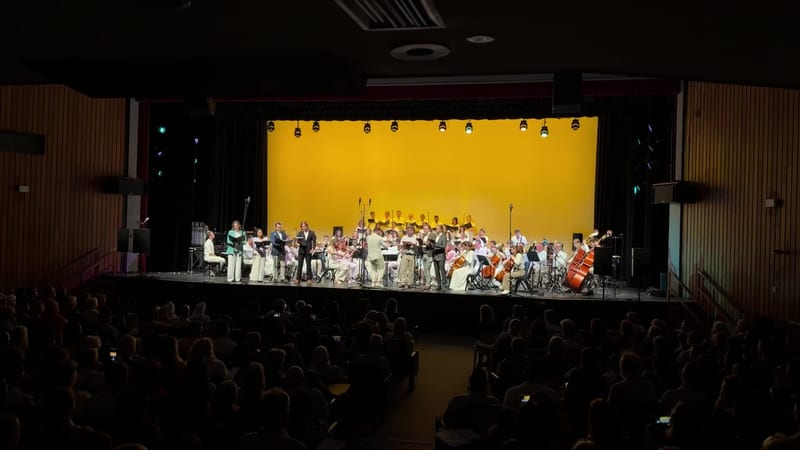 A full orchestra and choir perform together on stage under bright yellow lighting with an engaged audience in the foreground.