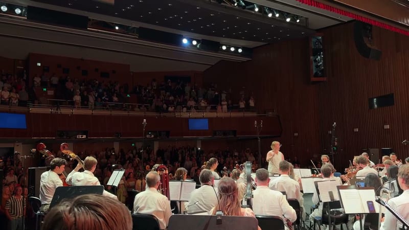 A conductor leads the orchestra during a performance in a red-walled concert hall with audience members visible in the balcony.