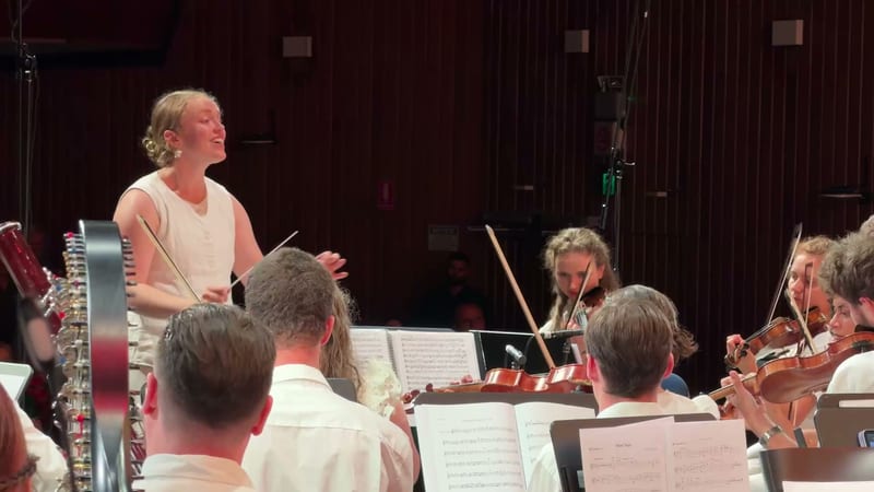 A conductor leads an orchestral rehearsal with string players seated with sheet music in a professional concert hall.