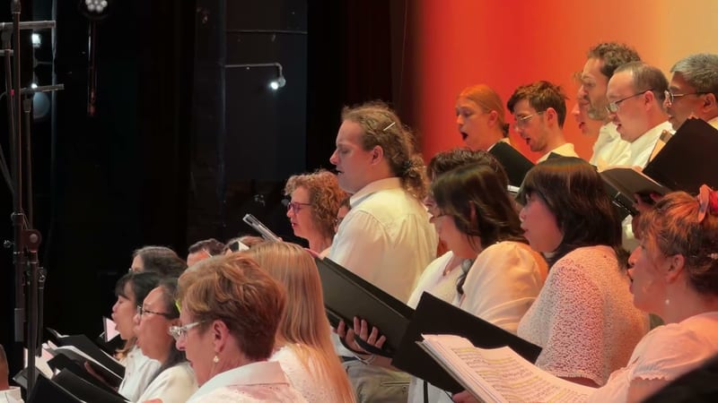 A choir of approximately twenty singers, mostly women and some men, perform together while holding sheet music under warm stage lighting.