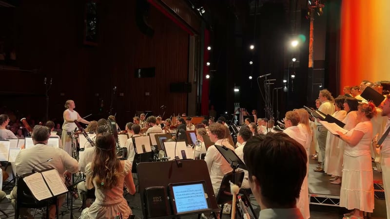 A conductor leads a large orchestra and choir ensemble during a performance in a professional concert venue with dramatic stage lighting.