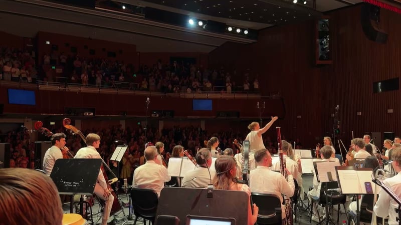 A conductor leads the orchestra during a performance in a concert hall with audience members visible in the balcony seating.