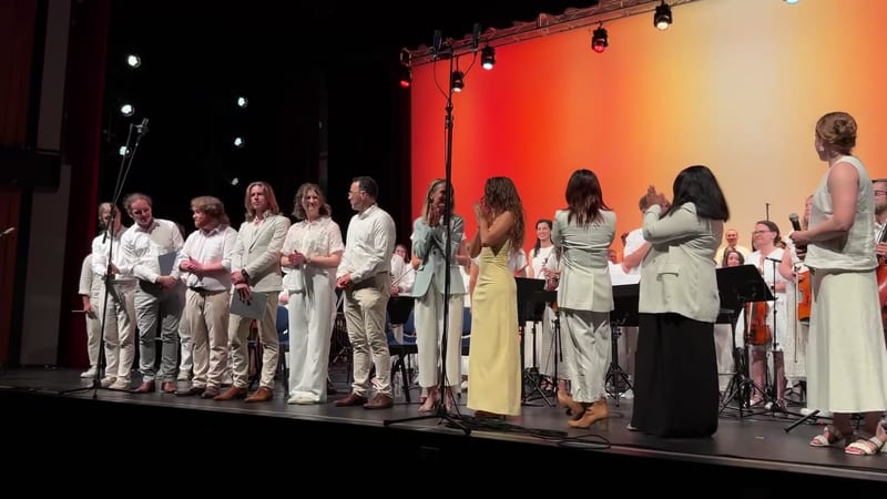 A choir and orchestra ensemble stand together on stage during a performance, with warm orange and red lighting illuminating the backdrop.