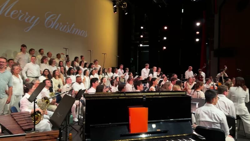 A large choir in white clothing performs alongside orchestral musicians with a grand piano and brass instruments on stage during a Christmas concert.