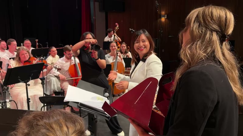 Orchestra members and conductor interact during a rehearsal or performance in an elegant concert hall with warm lighting.