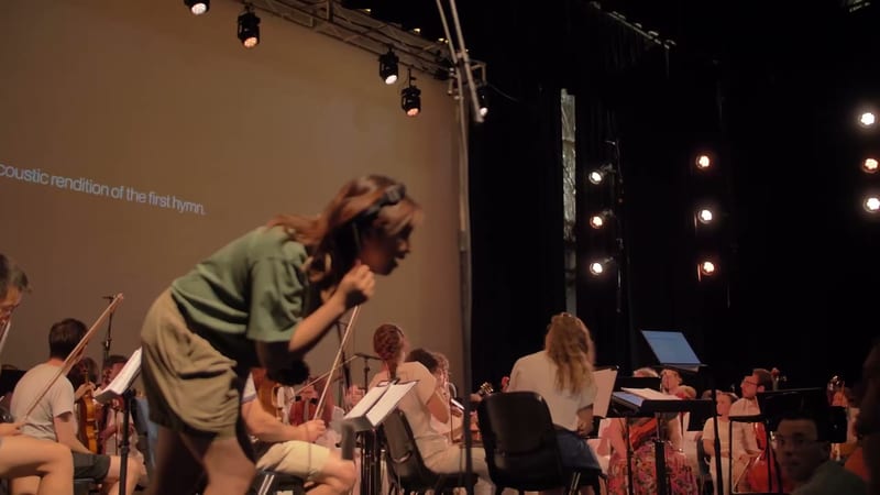 A conductor leads an orchestra during a performance, with musicians holding sheet music and instruments visible in the ensemble.
