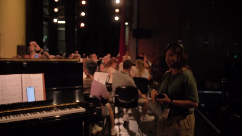 A conductor leads a rehearsal or performance with orchestra and choir members gathered in an auditorium with stage lighting.