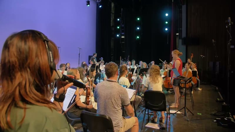 Orchestra members perform during a rehearsal or concert on stage with purple ambient lighting.