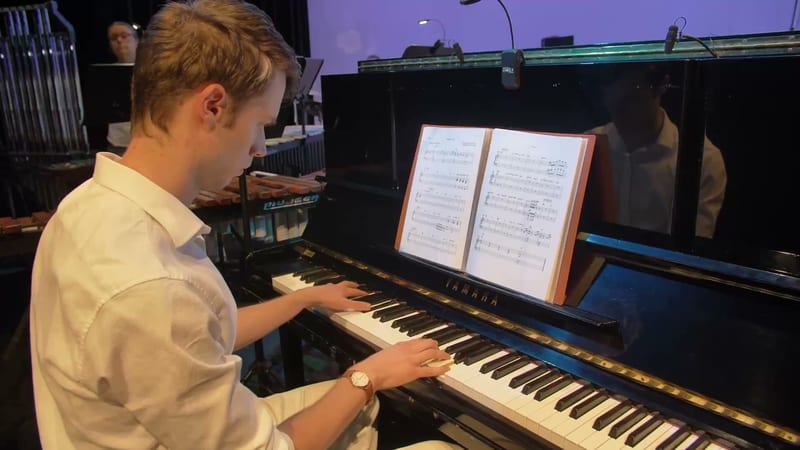 A young pianist performs at a grand piano while reading sheet music during a rehearsal or performance.