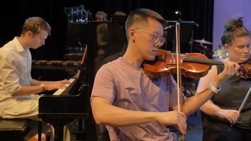 Young violinist in glasses performs with pianist and other string players during an orchestral rehearsal or performance.