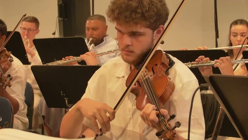 A violinist in white shirt concentrates while performing with orchestra members reading from sheet music stands.