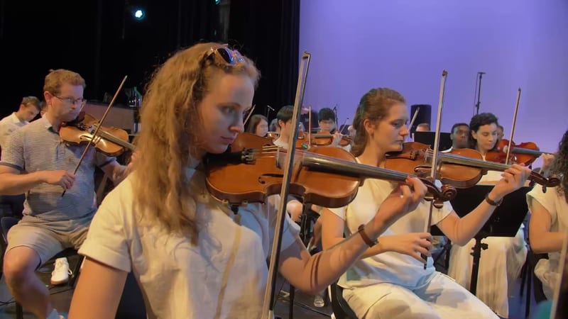 String section musicians perform together during an orchestral rehearsal or concert under purple stage lighting.