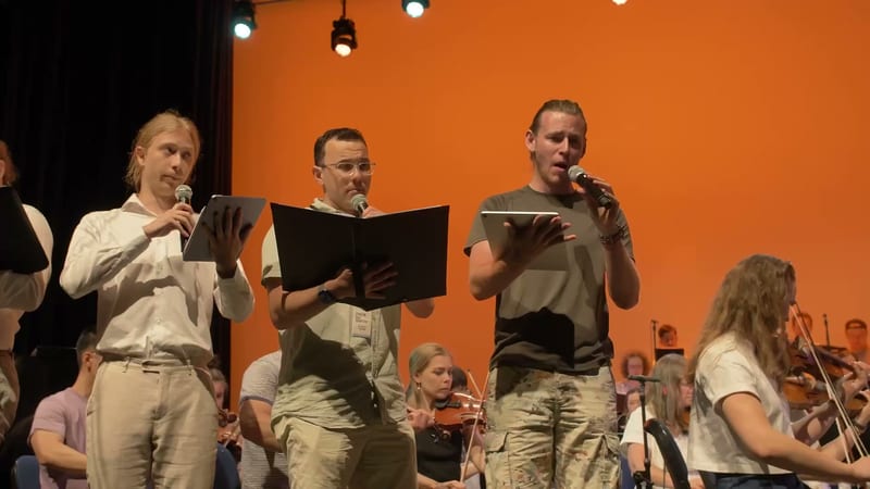 Four performers hold scripts and sing into microphones on stage with an orchestra and orange backdrop behind them.