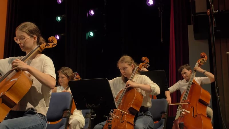 Young cellists perform together on stage during an orchestral concert with stage lighting.