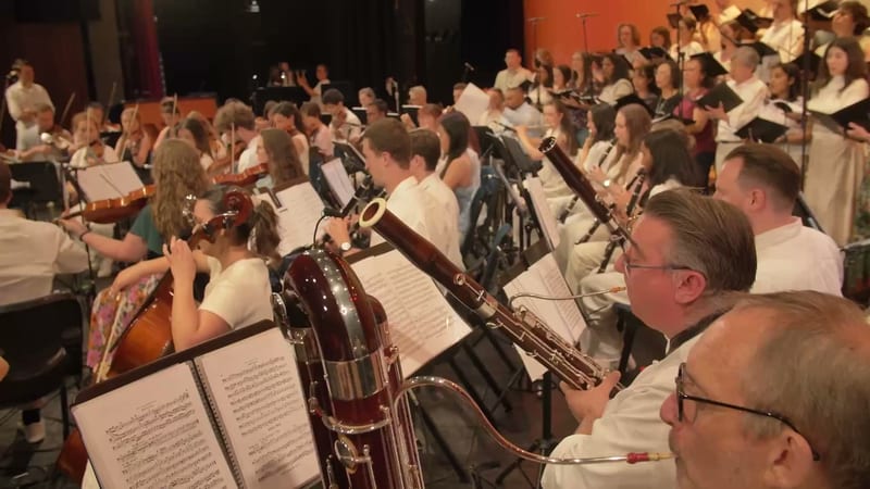 Orchestra musicians in white shirts perform together with sheet music and instruments during a rehearsal or concert.