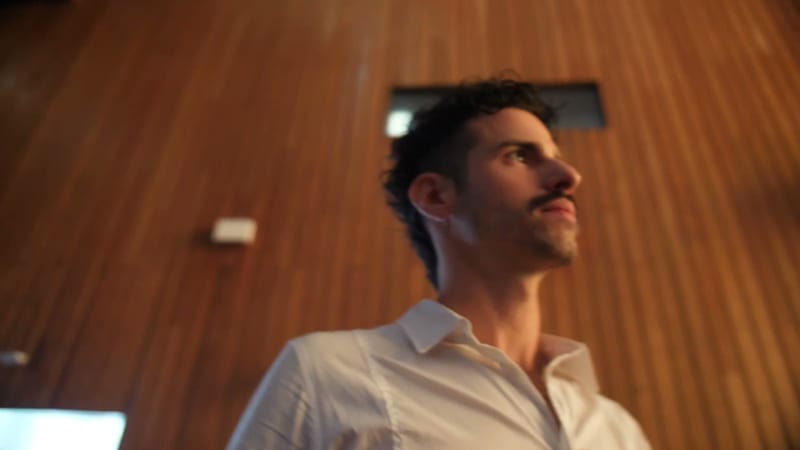 A man in a light blue shirt looks upward thoughtfully in an indoor venue with warm wood paneling.