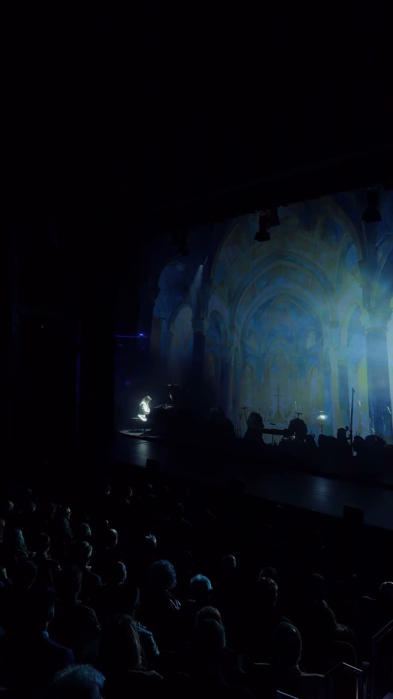 A performer plays an illuminated piano on stage with a vibrant backdrop.